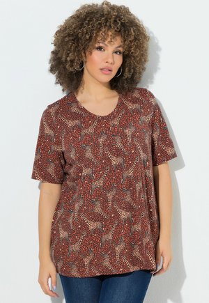 Vrouw met krullend haar die creolen draagt en een roestkleurig T-shirt met lichtbruine girafpatronen, gecombineerd met donkerblauwe spijkerbroek.