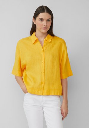 Chemise en lin jaune à manches courtes avec un col et une fermeture à boutons. Le tissu présente une finition texturée et une coupe décontractée et ample.
