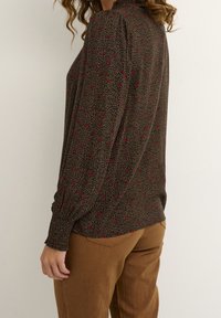 Woman in brown pants and long-sleeve black blouse with small red floral pattern, standing sideways against a plain background.