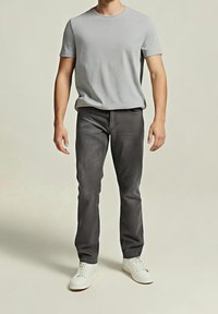 Homme debout portant un t-shirt gris clair, un pantalon gris foncé et des baskets blanches sur un fond uni. Haut du visage non visible.