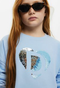 Sudadera de color azul claro con un gran diseño de lentejuelas en plata y azul. Modelo que lleva gafas de sol cat-eye negras y tiene el cabello largo y rojo.