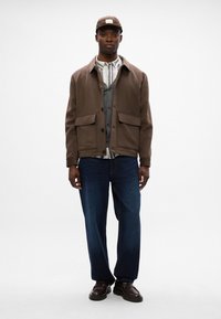 Giubbotto di cotone marrone con grandi tasche frontali, indossato sopra un gilet grigio e una camicia a scacchi, abbinato a jeans larghi blu scuro e scarpe marroni.
