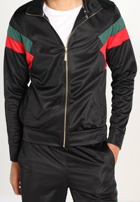 Svart zip-up jacka med gröna och röda accenter på axlarna; tillverkad av slät material; har en guldfärgad dragkedja och ribbad mudd.