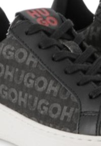 Baskets noires avec une tige texturée arborant un logo "Hugo" répété, des lacets noirs et une semelle en caoutchouc blanche. Comprend des accents de logo rouges.