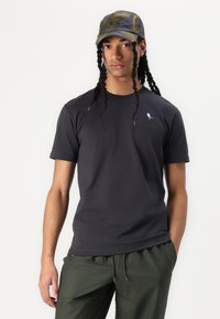 Dunkelgraues, kurzes T-Shirt mit einem kleinen blauen Vogelstick. Kombiniert mit olivgrünen, leichten, elastischen Hosen mit elastischem Bund. Lässiger Stil.
