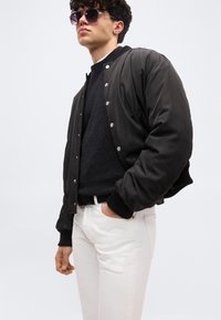 Νεαρός άνδρας που φοράει μαύρο μπουφάν τύπου bomber, σκούφο πουλόβερ, λευκό παντελόνι και σκούρα γυαλιά ηλίου, με το ένα χέρι στην τσέπη, μπροστά σε απλό φόντο.