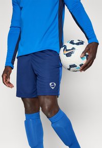 Μπλε μακρυμάνικο μπλουζάκι με ναυτά ποδιές στην πλευρά, ναυτικές κοντές βερμούδες με λευκό λογότυπο Nike, σε συνδυασμό με μπλε κάλτσες μέχρι το γόνατο και μια σχεδιασμένη μπάλα ποδοσφαίρου.