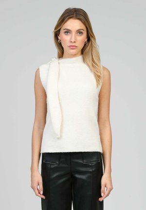 Femme aux longs cheveux blonds portant un haut en maille blanc sans manches avec un détail en écharpe et un pantalon en cuir noir, faisant face à l'avant sur un fond gris.