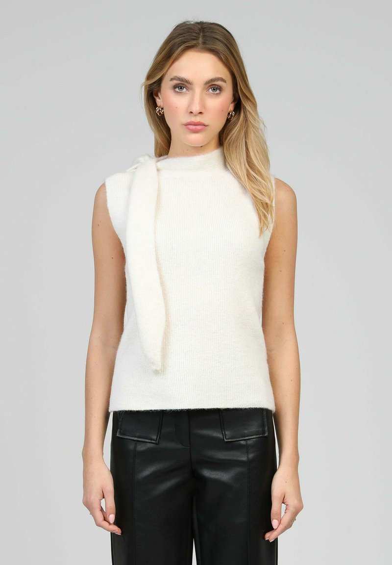 Femme aux longs cheveux blonds portant un haut en maille blanc sans manches avec un détail en écharpe et un pantalon en cuir noir, faisant face à l'avant sur un fond gris.