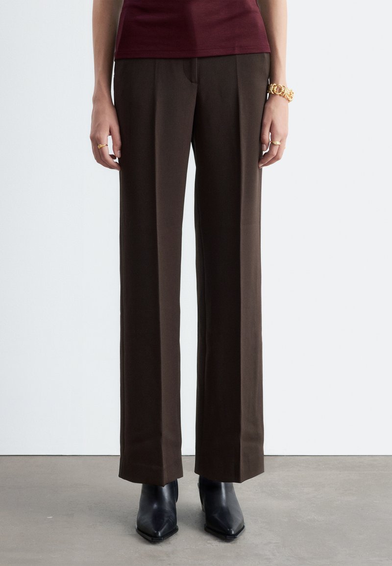 Personne portant un pantalon droit marron foncé, des bottes noires pointues, un haut bordeaux et des bijoux en or, debout sur un sol gris.