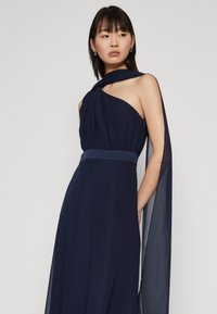 Robe de soirée navy avec un design à épaule unique, en tissu chiffon fluide, taille cintrée et accent d'une longue écharpe drapée.