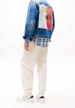 Person trägt hellbeige Jogginghosen, weiße Sneaker, blau kariertes Hemd unter einer Jeansjacke mit einem rot-weißen Aufnäher und dem Schriftzug "TIMMY" auf dem Rücken.