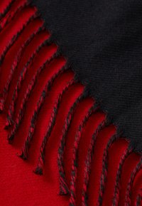 Cachecol preto e vermelho com detalhes em franjas. O tecido preto é suave, contrastando com o corpo vermelho vibrante e liso, apresentando franjas torcidas.