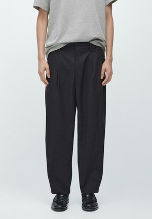 Person trägt lockere schwarze plissierte Hosen, schwarze Loafers und ein graues Kurzarmhemd und steht vor einem schlichten hellen Hintergrund.