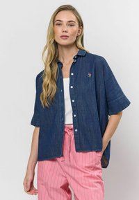 Džínová košeľa s krátkymi rukávmi, zapínaním na gombíky a vyšitým detailom. Tkanina je tmavomodrá a má hladkú textúru.