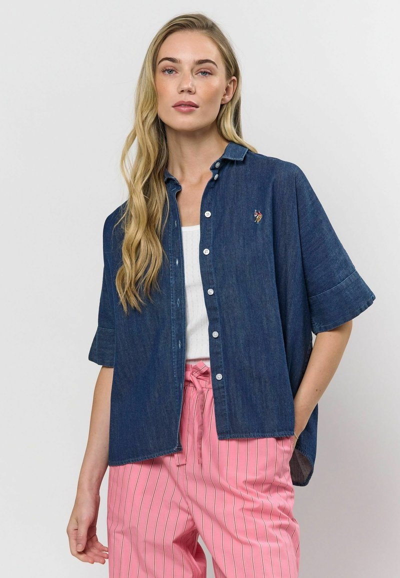Džínová košeľa s krátkymi rukávmi, zapínaním na gombíky a vyšitým detailom. Tkanina je tmavomodrá a má hladkú textúru.