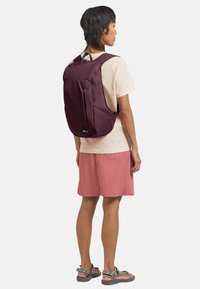 Persona in una t-shirt beige, pantaloni corti rosa e sandali fantasia, con uno zaino bordeaux, in piedi di lato contro uno sfondo semplice.
