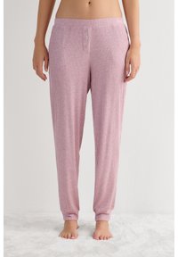 Vrouw die hoge taille roze geribbelde loungebroek met knoopdetails draagt, blootsvoets staand op een zacht wit tapijt tegen een effen muur.
