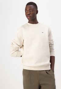 Lys beige genser med ribbet rund hals og mansjetter, med en liten tri-farge logo på brystet. Kombinert med olivengrønne bukser.