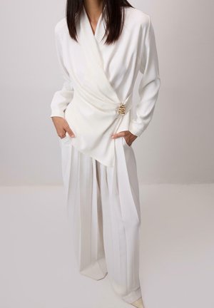 Femme portant un élégant chemisier blanc cache-cœur avec fermoir doré et un pantalon blanc plissé à jambes larges, debout les mains dans les poches.