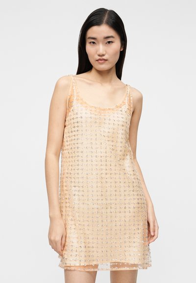 Jovem mulher vestindo um mini vestido de alças cor pêssego com aplicações de estrelas prateadas, a posar contra um fundo claro e simples.