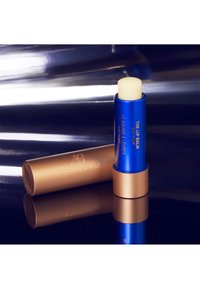 Blaues und goldenes Lippenbalsam-Röhrchen mit der Aufschrift "The Lip Balm" neben seiner goldenen Kappe mit "Augustinus Bader" auf einer reflektierenden dunklen Oberfläche.