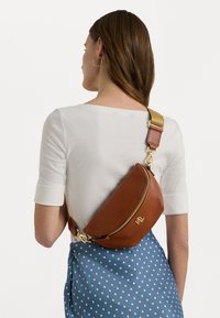 Brun lærveske med en buet form, gullhardware og en avtakbar, beige stropp. Båren over en hvit topp og blå prikkete skjørt.