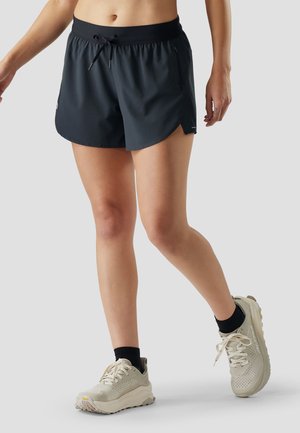 Kvinde iført sorte sportsshorts, sorte ankelstrømper og lysebeige løbesko, stående med det ene ben let bøjet.