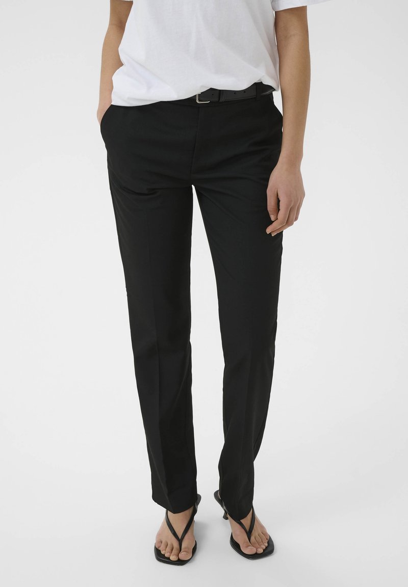 Personne portant un pantalon noir slim, une ceinture noire, une chemise blanche rentrée, et des sandales à talons noires ouvertes au bout, debout les mains dans les poches.