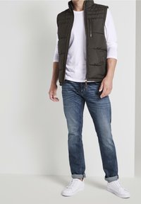 Gepolsterte, ärmellose Weste in Dunkelgrau mit Reißverschlusstasche, getragen über einem weißen Langarmshirt. Kombiniert mit blauen Jeans und weißen Turnschuhen.