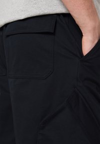 Main dans la poche d’un pantalon foncé avec une poche à rabat à l’arrière et une poche latérale inclinée, avec une partie d’un haut gris clair visible.