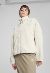 Giacca in pile bianca con colletto alto, chiusura con zip e un piccolo logo ricamato. Abbinata a pantaloni beige a coste.