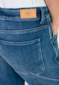 Jean en denim bleu avec une étiquette en cuir à la taille, comportant deux poches arrière et un motif de couture distinctif sur les bords des poches.