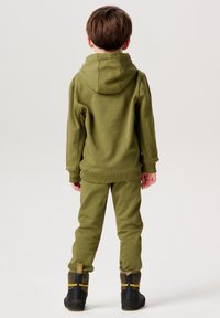 Olivgrüner Kapuzenpullover und passende Jogginghose aus weichem Stoff. Verfügt über gerippte Bündchen und eine entspannte Passform. Getragen mit schwarzen Stiefeln.