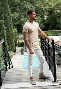 Chemise beige ajustée à manches courtes avec motifs géométriques, associée à un pantalon blanc slim et des mocassins gris, debout sur une terrasse en bois.