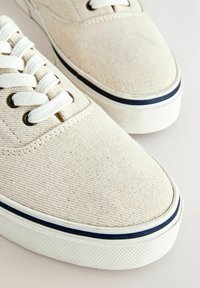 Baskets en toile beige avec lacets blancs, semelle en caoutchouc blanche et détail de bande noire sur la semelle intermédiaire.