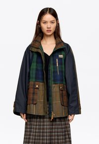 Multi-uzorčena jakna s kariranim in enotnim delom v zeleni, temno modri in rjavi barvi. Ima zadrgo spredaj, žepe in visok ovratnik.