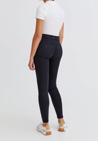 Jeans negros ajustados con una cintura alta, bolsillos traseros y una tela elástica. Combinados con una camiseta blanca ajustada y zapatillas de color claro.