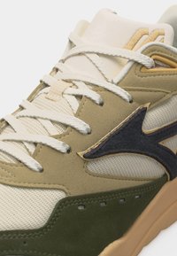 Beige og grønn joggesko med hvite lisser, paneler i mesh og semsket skinn, og en svart bølgeformet logo på siden.