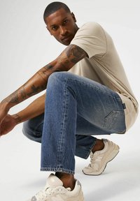 Camiseta de manga corta beige, pantalones vaqueros azul desgastado y zapatillas blancas. Tatuajes visibles en los brazos, postura de cuclillas, fondo neutro.
