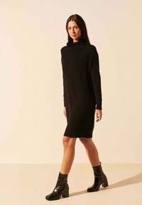 Robe-pull noire à col montant et manches longues. Confectionnée dans une matière douce, elle tombe au-dessus des genoux. Assortie à des bottines noires à talons.