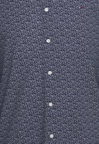 Chemise bleu marine avec motif floral blanc, boutons blancs sur le devant et un petit logo rouge, blanc et bleu près du coin supérieur droit.