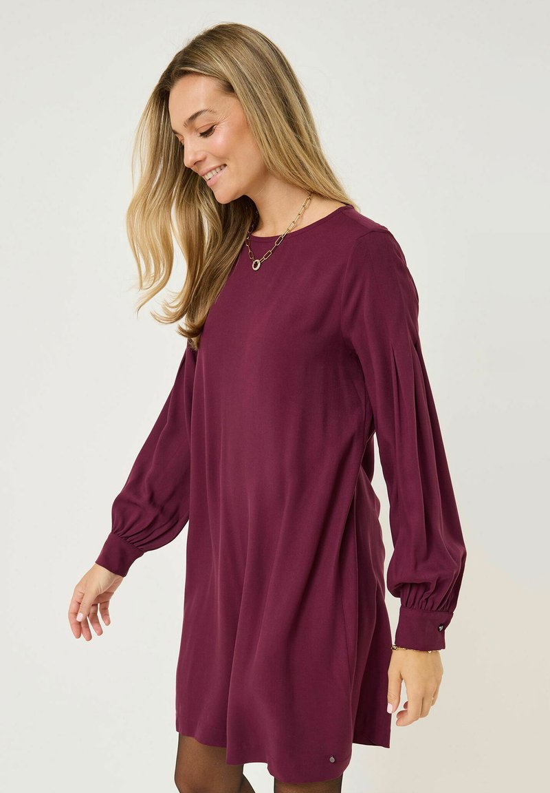 Abito bordeaux con maniche a sbuffo lunghe e scollatura rotonda. Tessuto liscio; vestibilità leggermente comoda. Piccolo dettaglio decorativo vicino all'orlo.