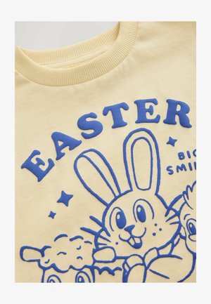 Sárga pulóver, kék nyomtatott grafikával, amelyen egy nyúl és karakterek láthatók, kiemelt "EASTER" felirattal és csillagokkal a mintán.