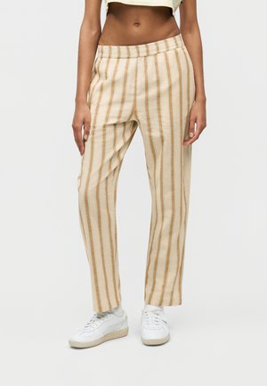 Femeie purtând pantaloni cu dungi verticale bej și albe, cu talie elastică, și adidași albi, stând în picioare pe fundal alb.