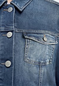 Κοντινό πλάνο στο μπροστινό μέρος μπλε τζιν μπουφάν που δείχνει κουμπωμένη τσέπη με καπάκι και ασημένια κουμπιά χαραγμένα με "Marc O'Polo est. 1967".