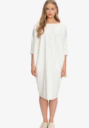 Kobieta o długich blond włosach ubrana w luźną, białą sukienkę do kolan z rękawami trzy czwarte i beżowe buty wsuwane.