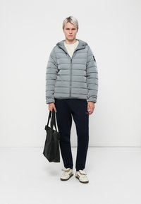 Lys grå pufferjakke med lynlås foran, hætte og horisontalt quiltet mønster. Båret over en cremefarvet sweatshirt og parret med marineblå bukser og en sort tote.