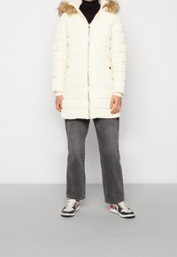 Chaqueta puffer crema con capucha forrada de pelo, diseño acolchado, cremallera frontal y bolsillos laterales, combinada con jeans grises y zapatillas negras.