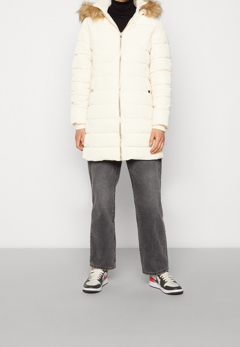 Chaqueta puffer crema con capucha forrada de pelo, diseño acolchado, cremallera frontal y bolsillos laterales, combinada con jeans grises y zapatillas negras.